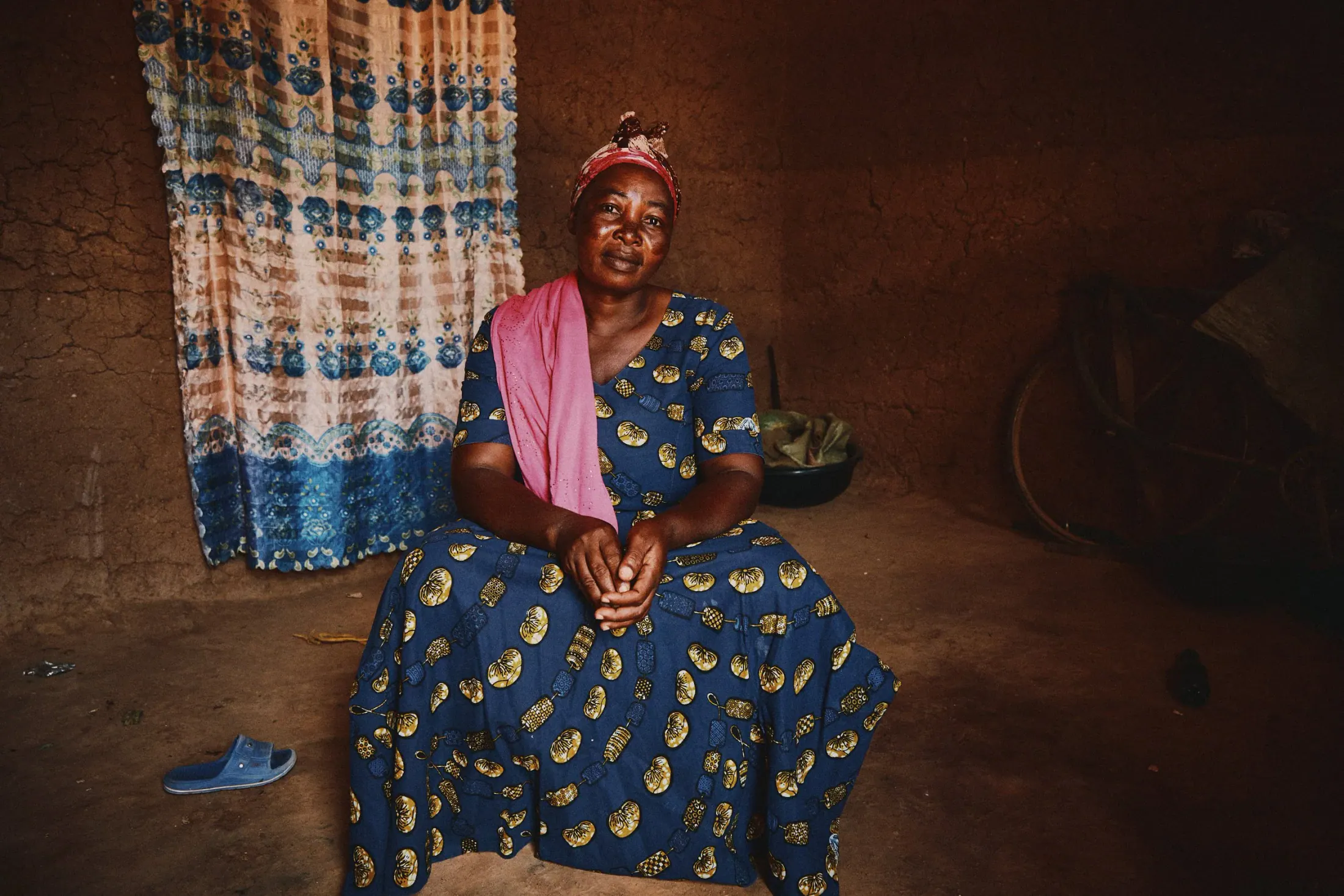 A woman in a blue dress sits in her home beside a patterned curtain