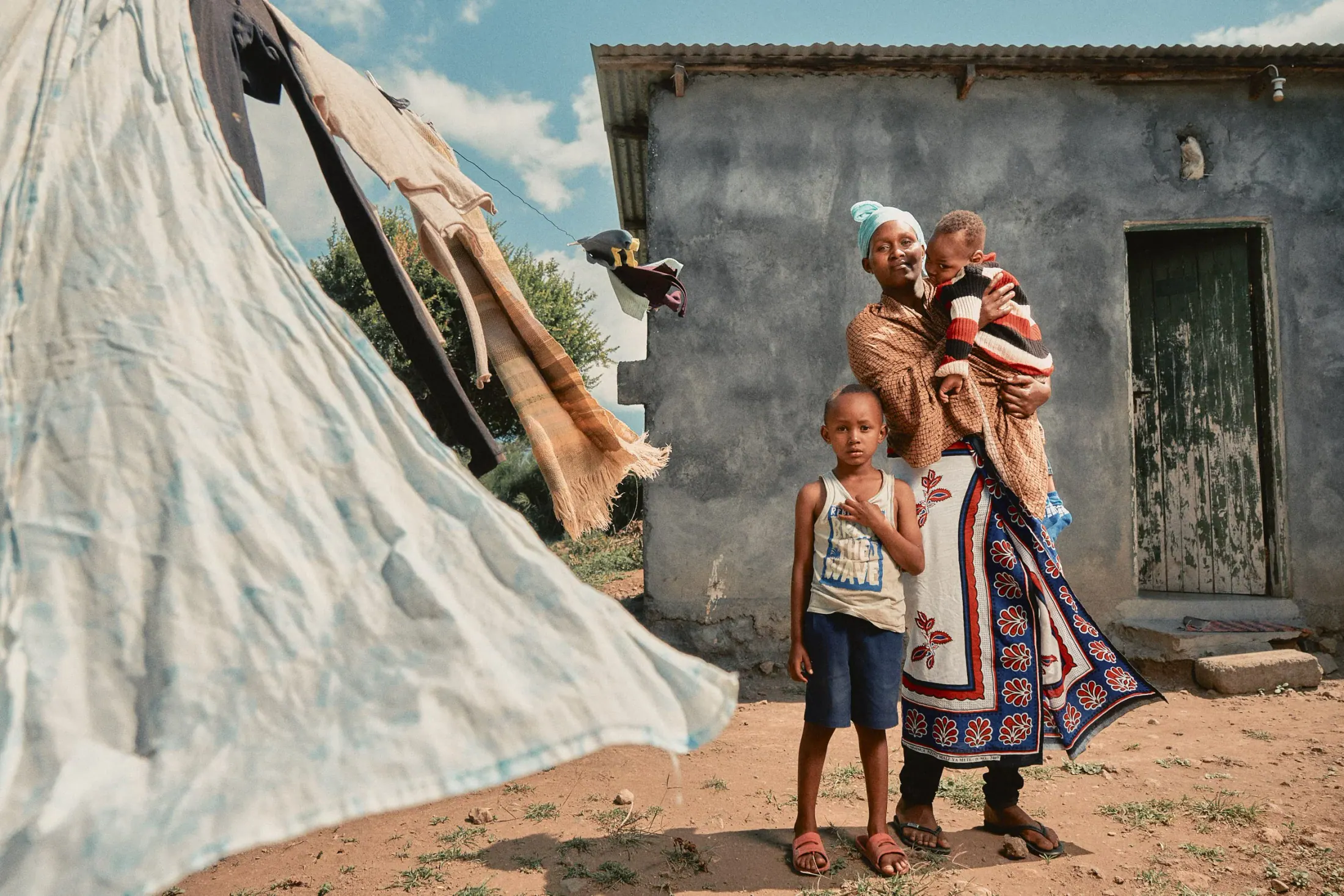 A mother holds her baby beside her daughter outside their house, washing drying in the wind