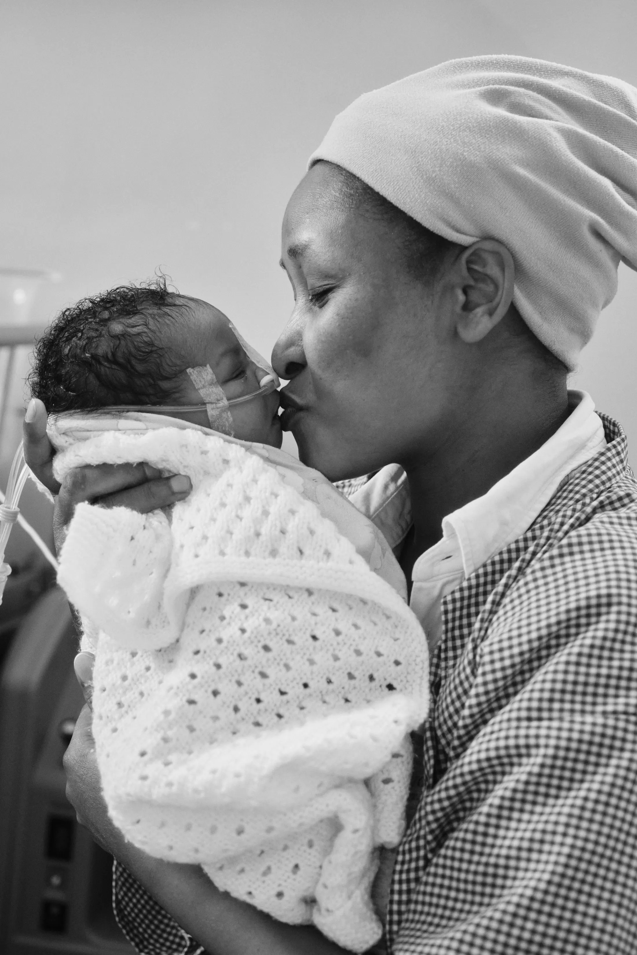 A mother kisses her newborn, swaddled in a white blanket (black and white)