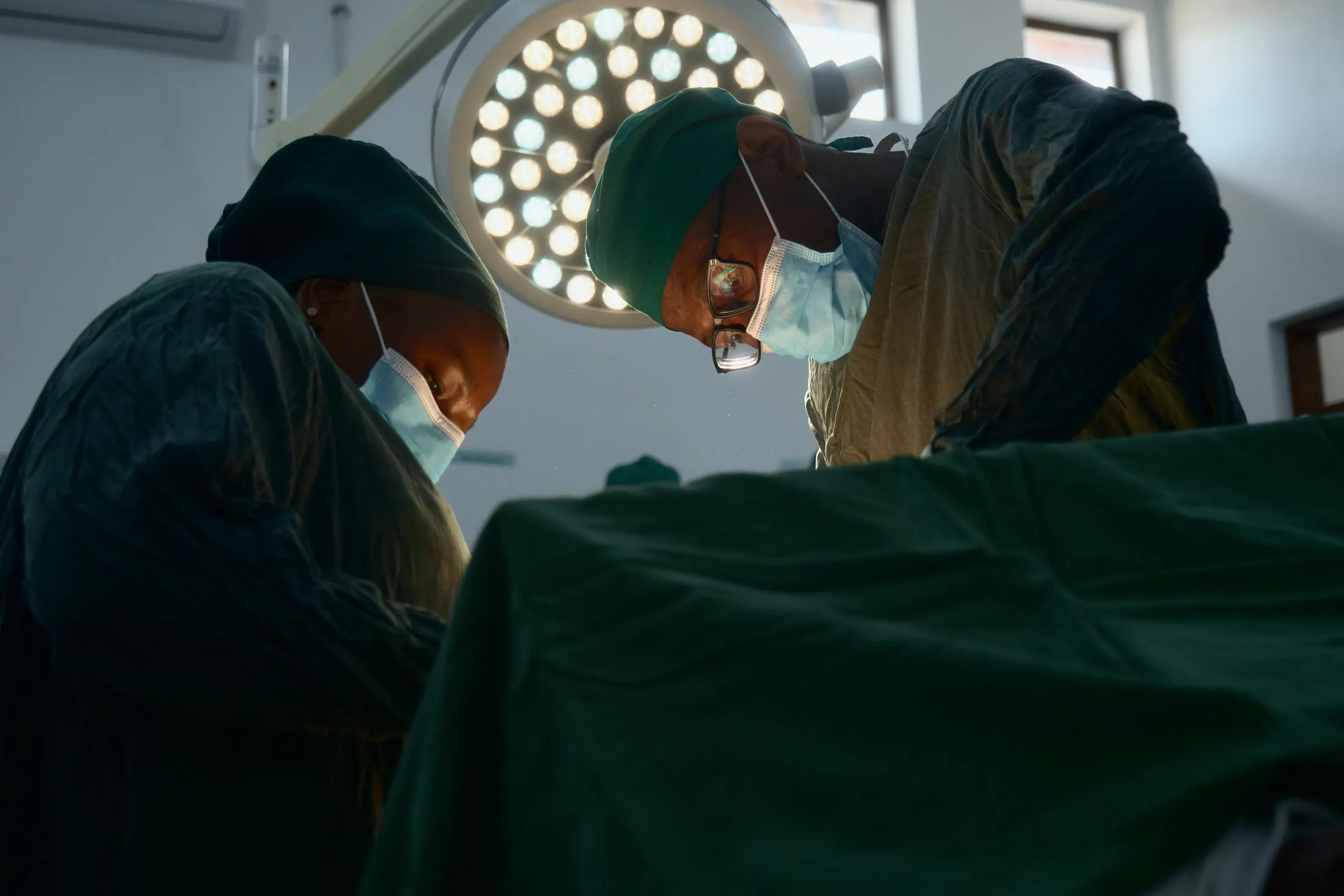 Two surgeons in masks and caps lean in under the operating lamp