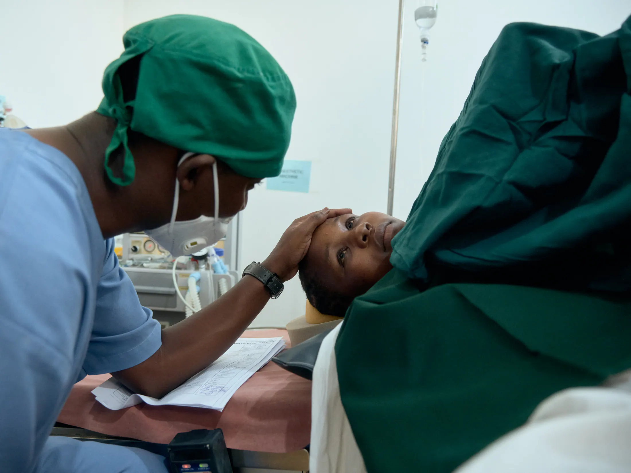 A nurse in a green cap rests her hand on a patient’s head in theatre
