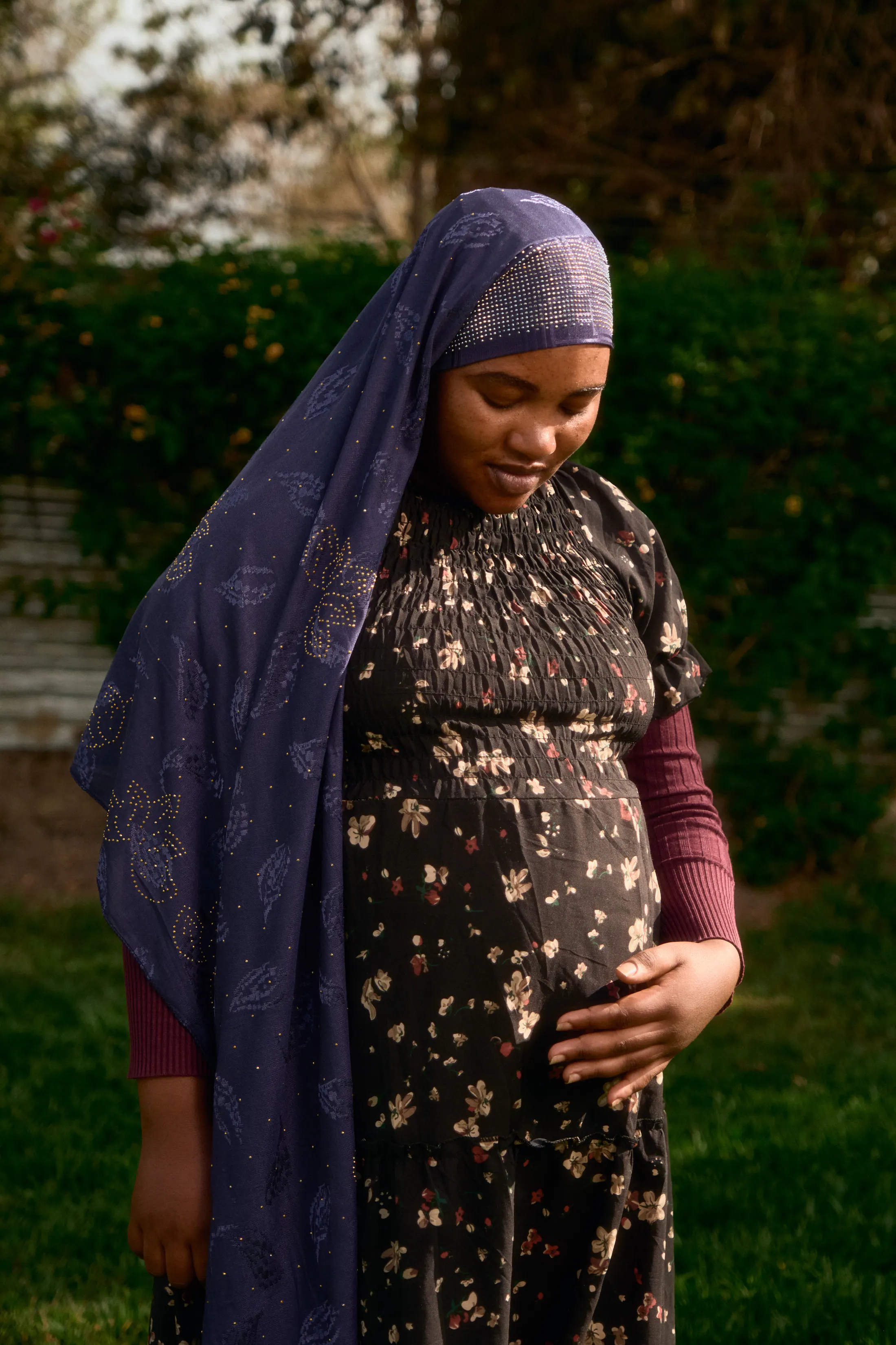 A pregnant woman in a purple headscarf stands in the garden, hand on her belly