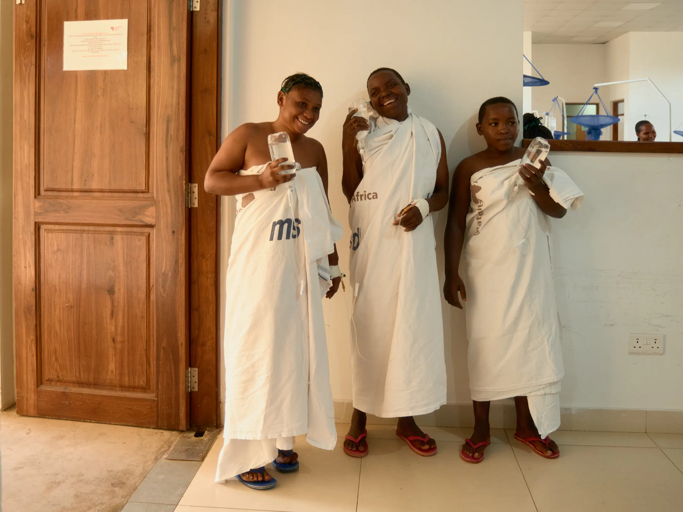 Three women in hospital gowns stand laughing in a ward doorway