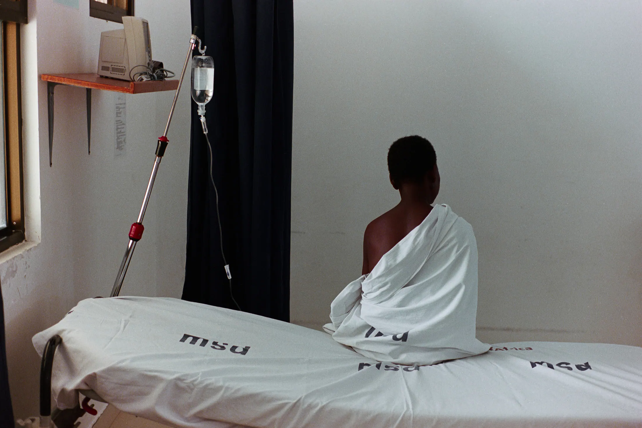 A woman wrapped in a sheet sits on the edge of a ward bed, facing the wall (35mm film)