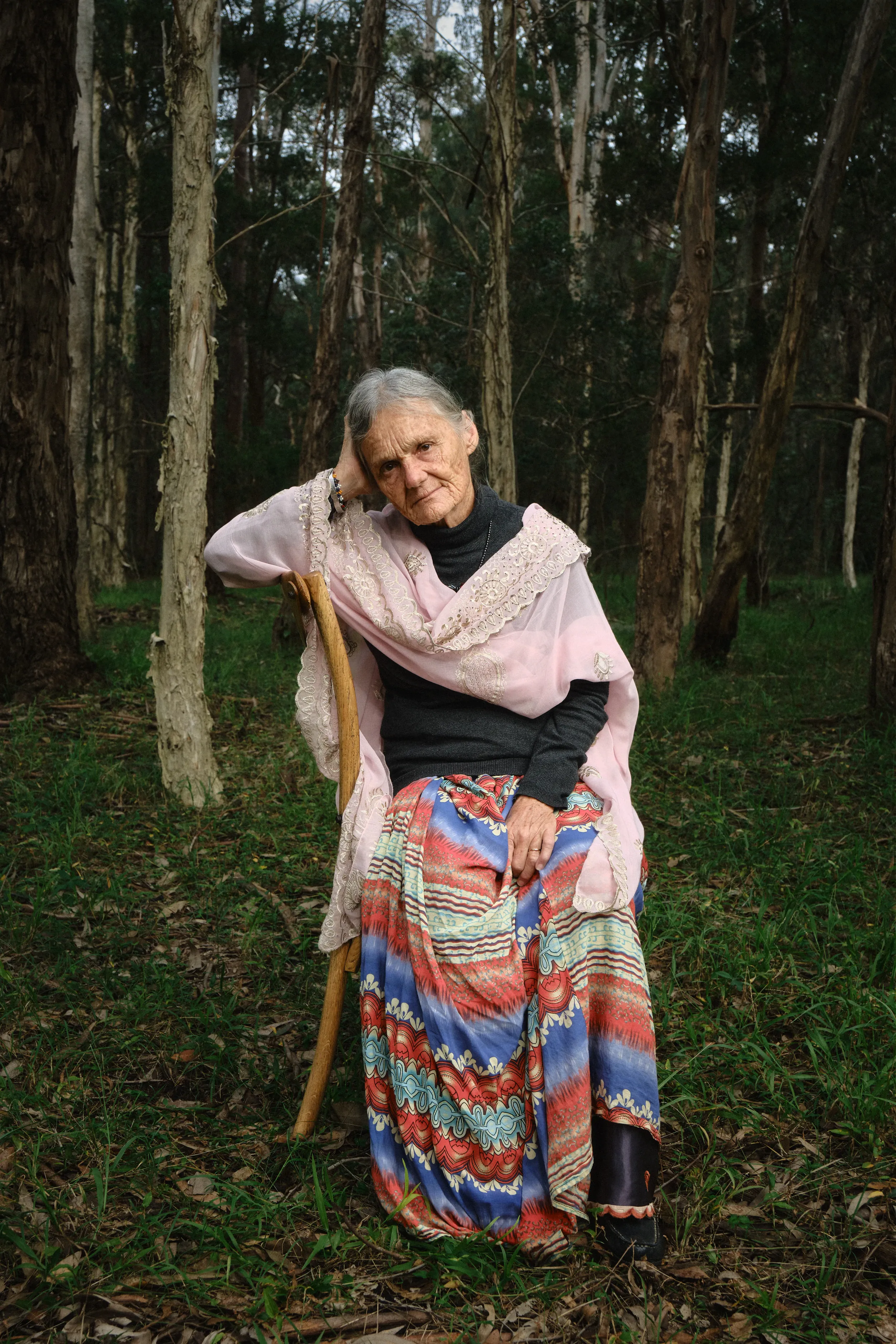 Valerie seated on a bentwood chair in the forest, one hand raised to her head