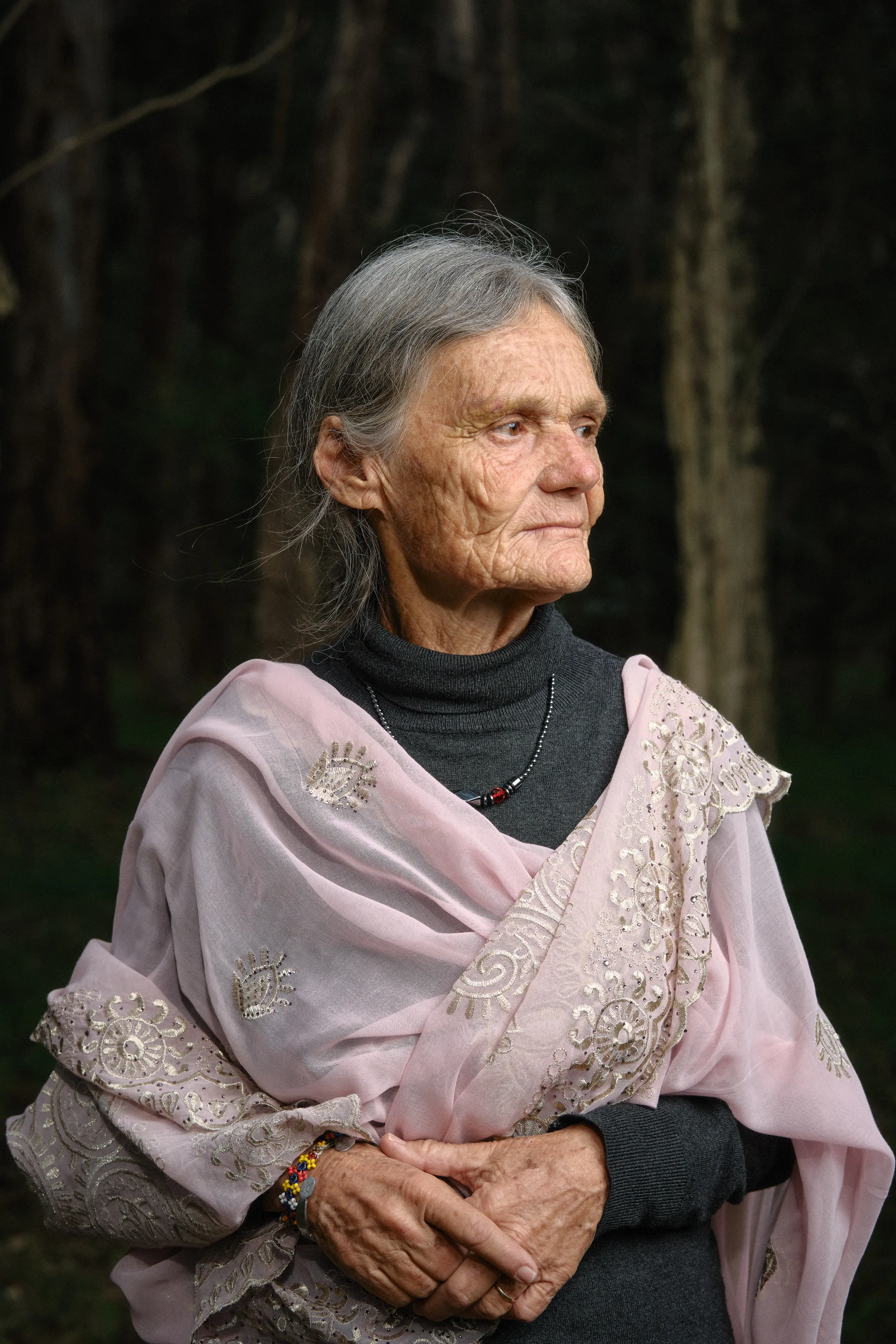 Valerie looks off to the side in soft forest light, hands folded over her shawl