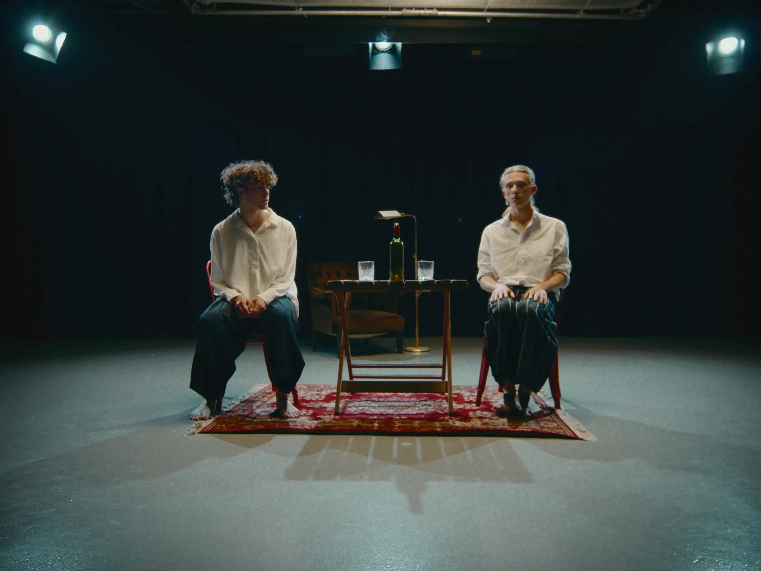 Two men in white shirts sit either side of a small table on a rug, lit by stage lights