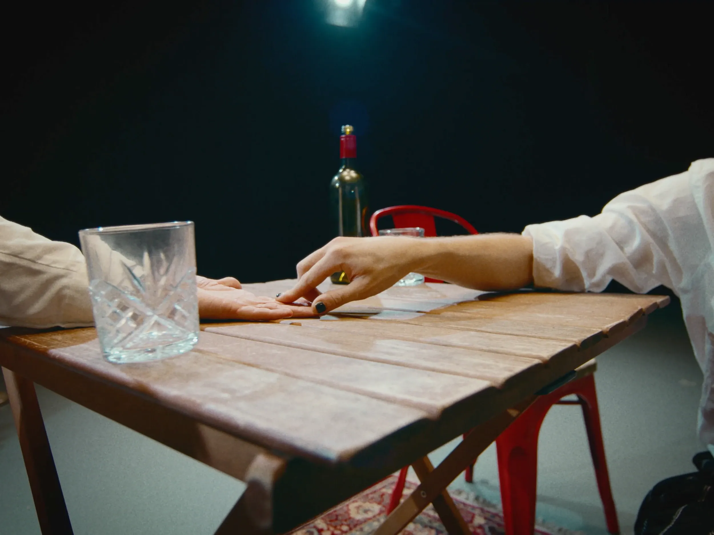 A hand rests beside a glass on the table, a rose in a bottle behind