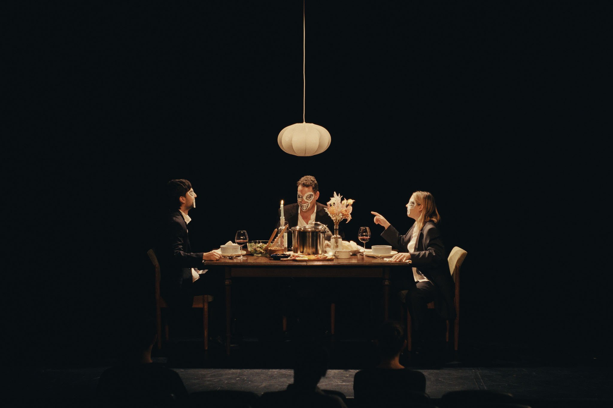 Three figures at a dinner table under a paper lantern on a black stage