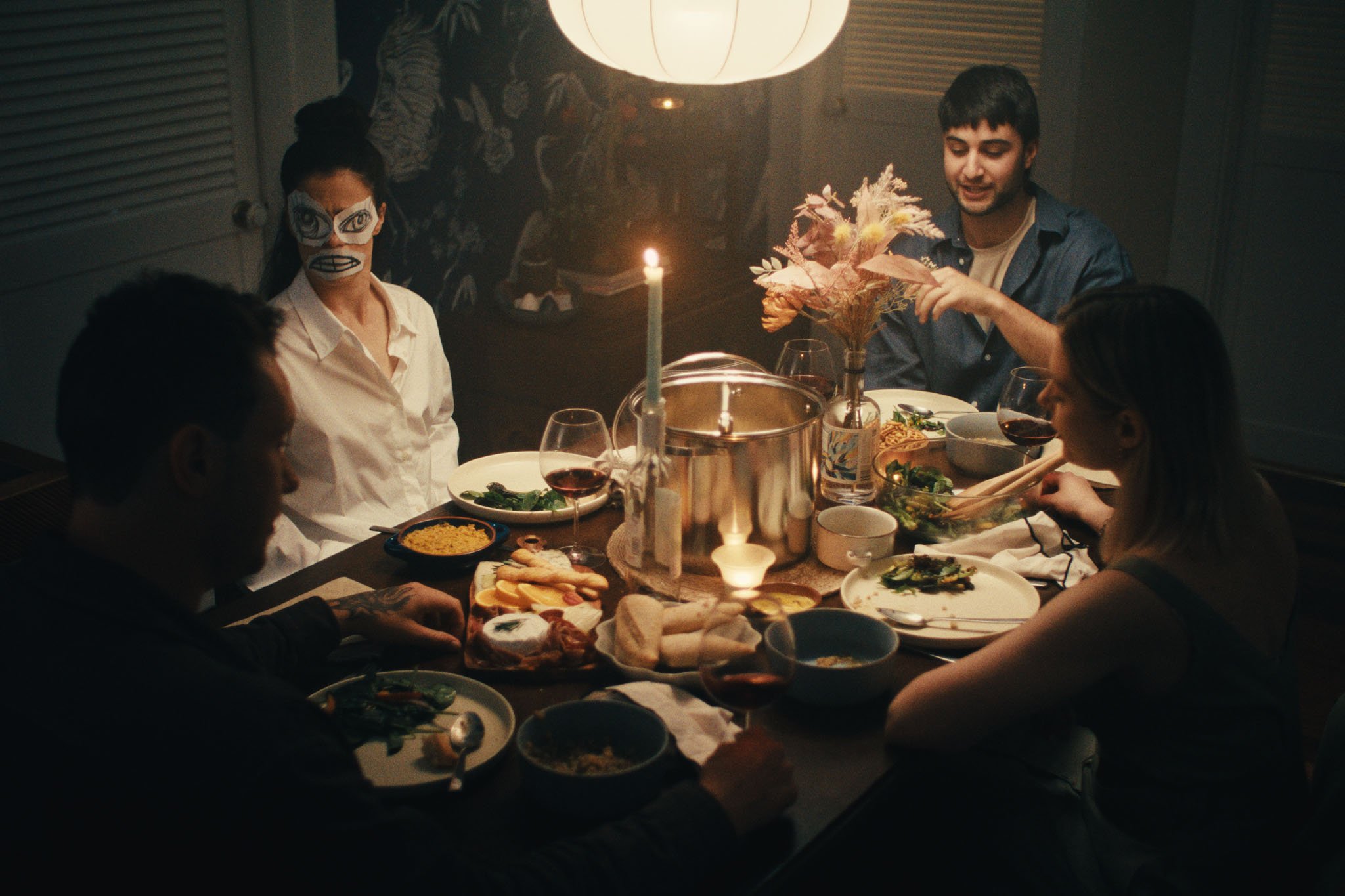 A candlelit family dinner; a woman in a paper mask sits among the guests