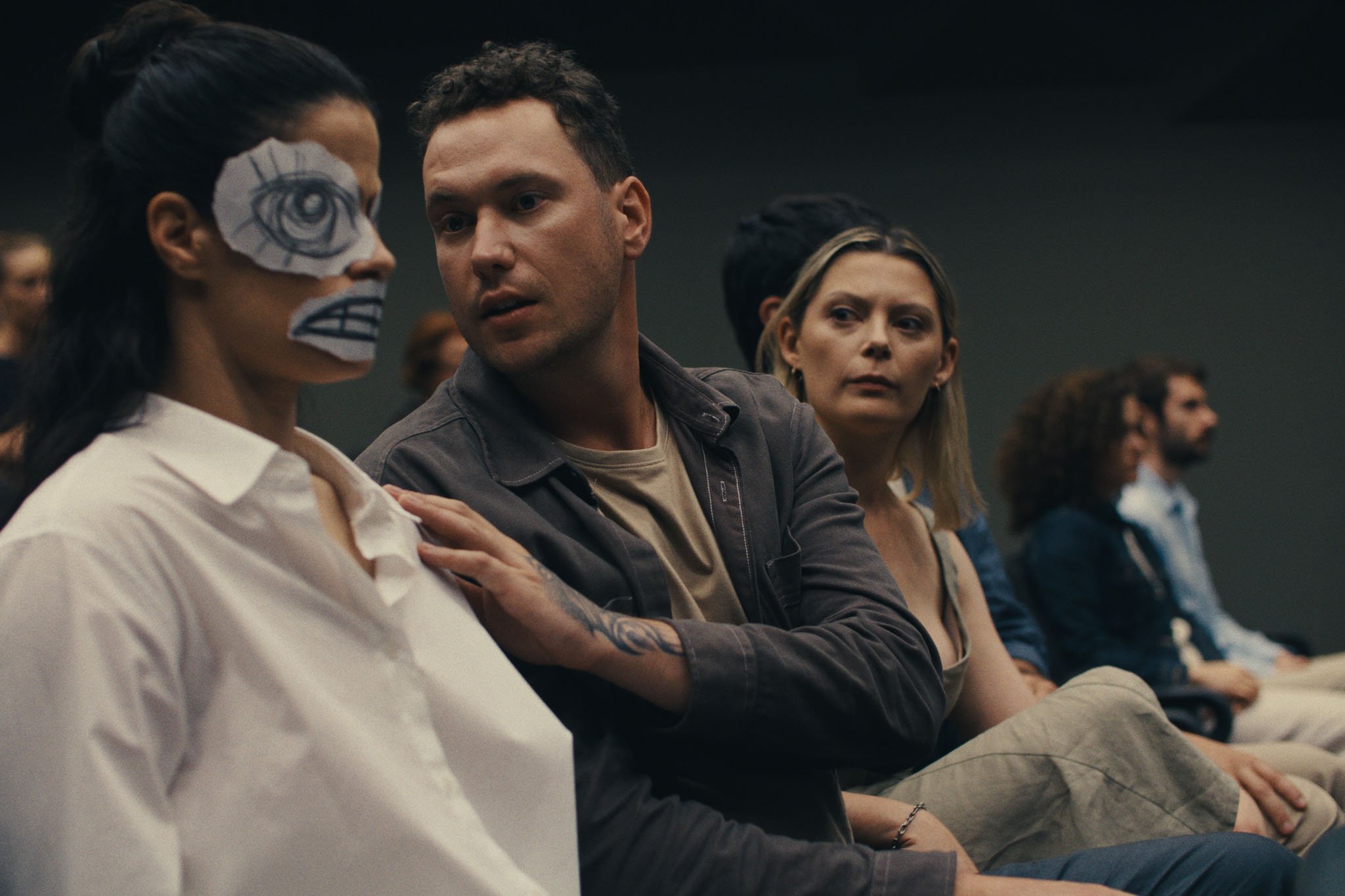 A woman in a paper eye-and-teeth mask sits in a crowd as a man touches her shoulder