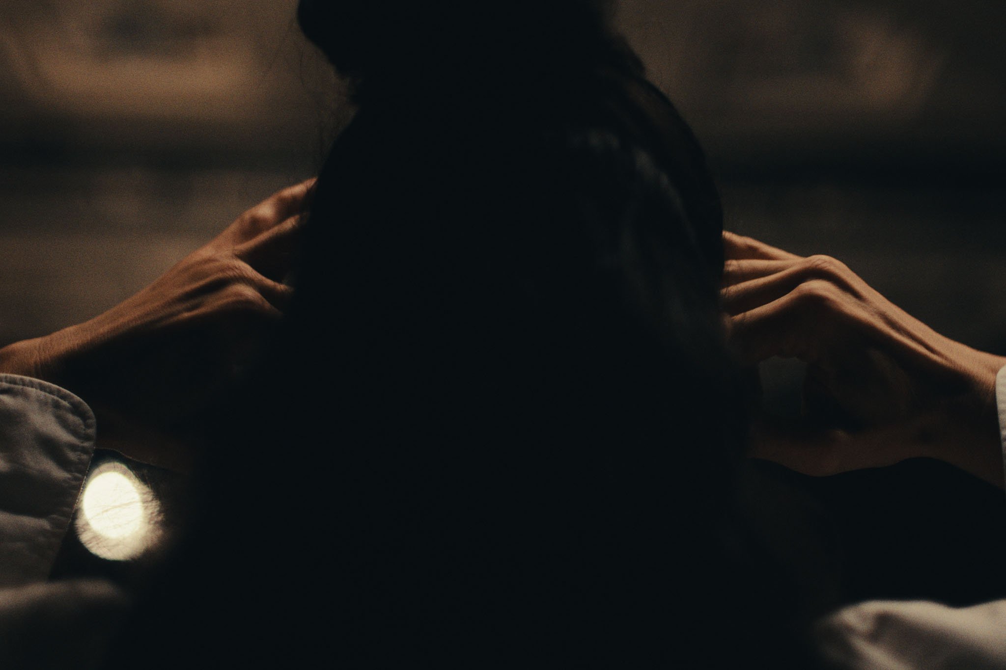 Hands reach toward a figure's shoulders in warm low light