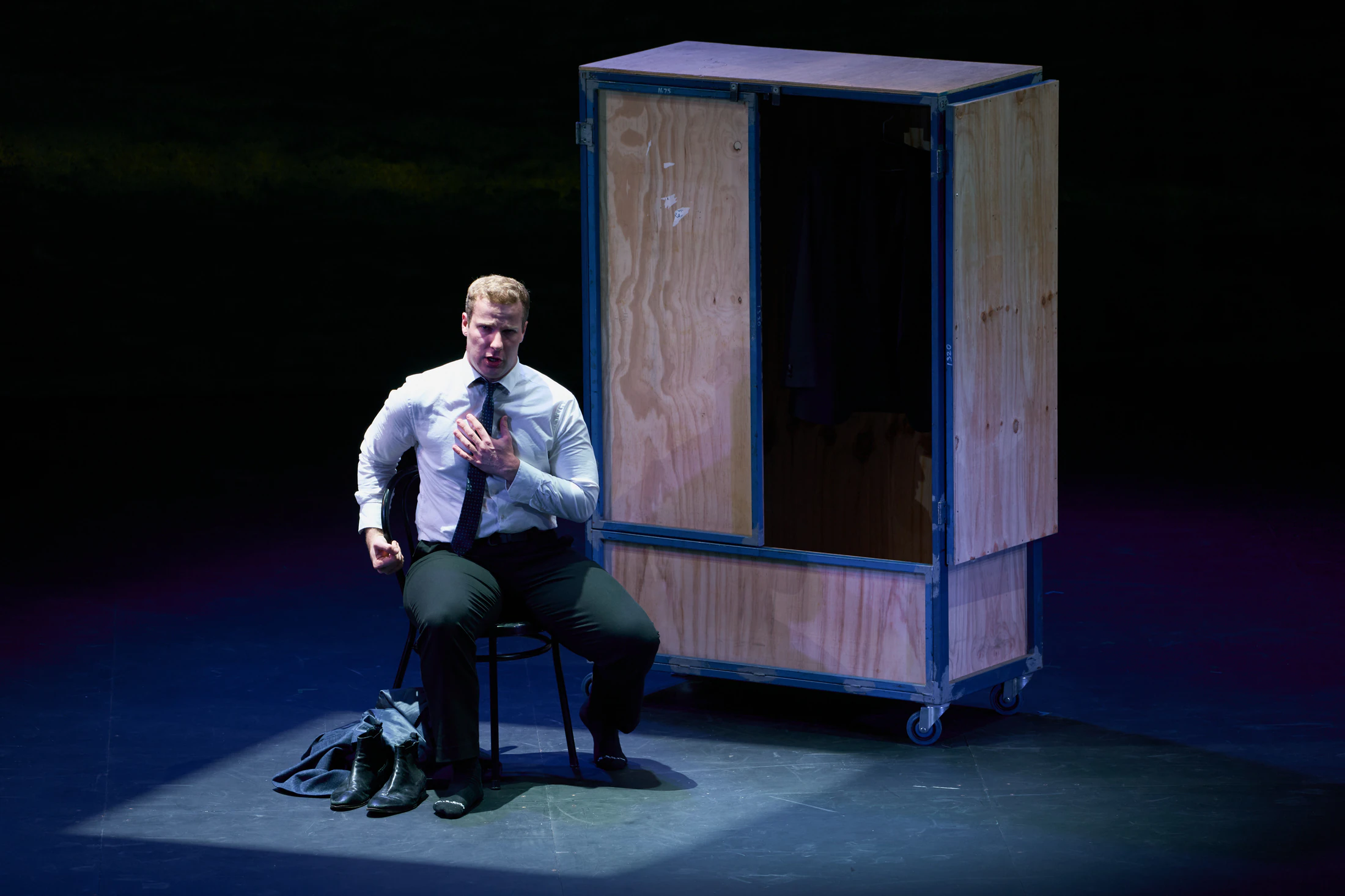 A singer sits alone in a pool of light beside a wooden wardrobe