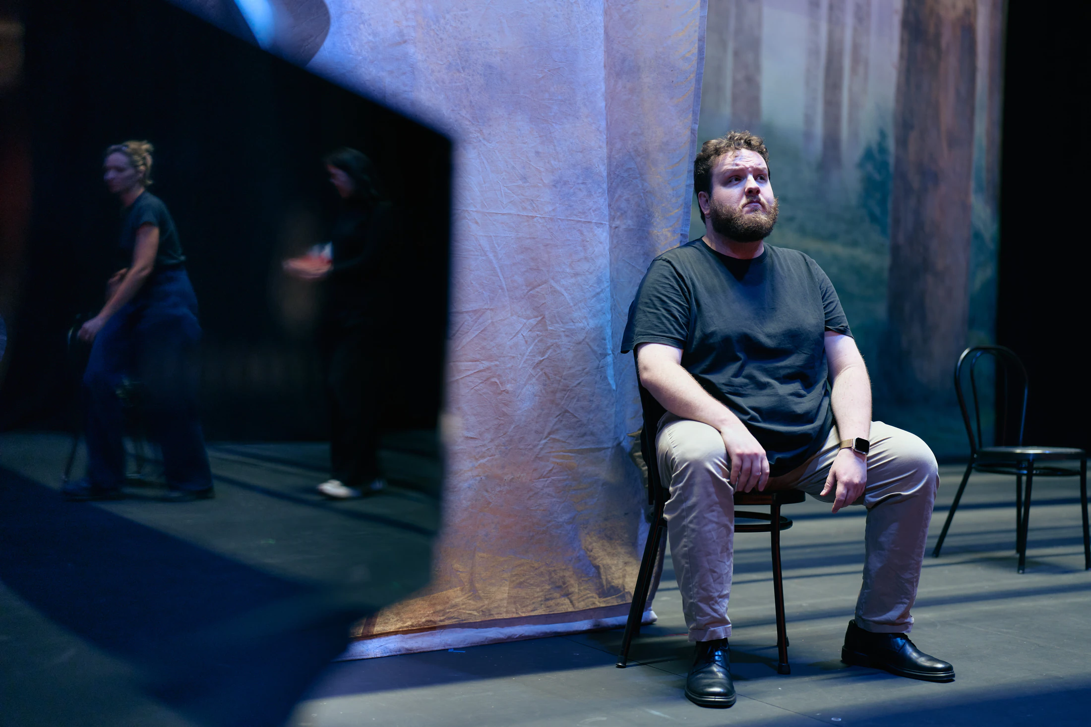 A singer sits on a chair on the dim stage, a painted forest behind