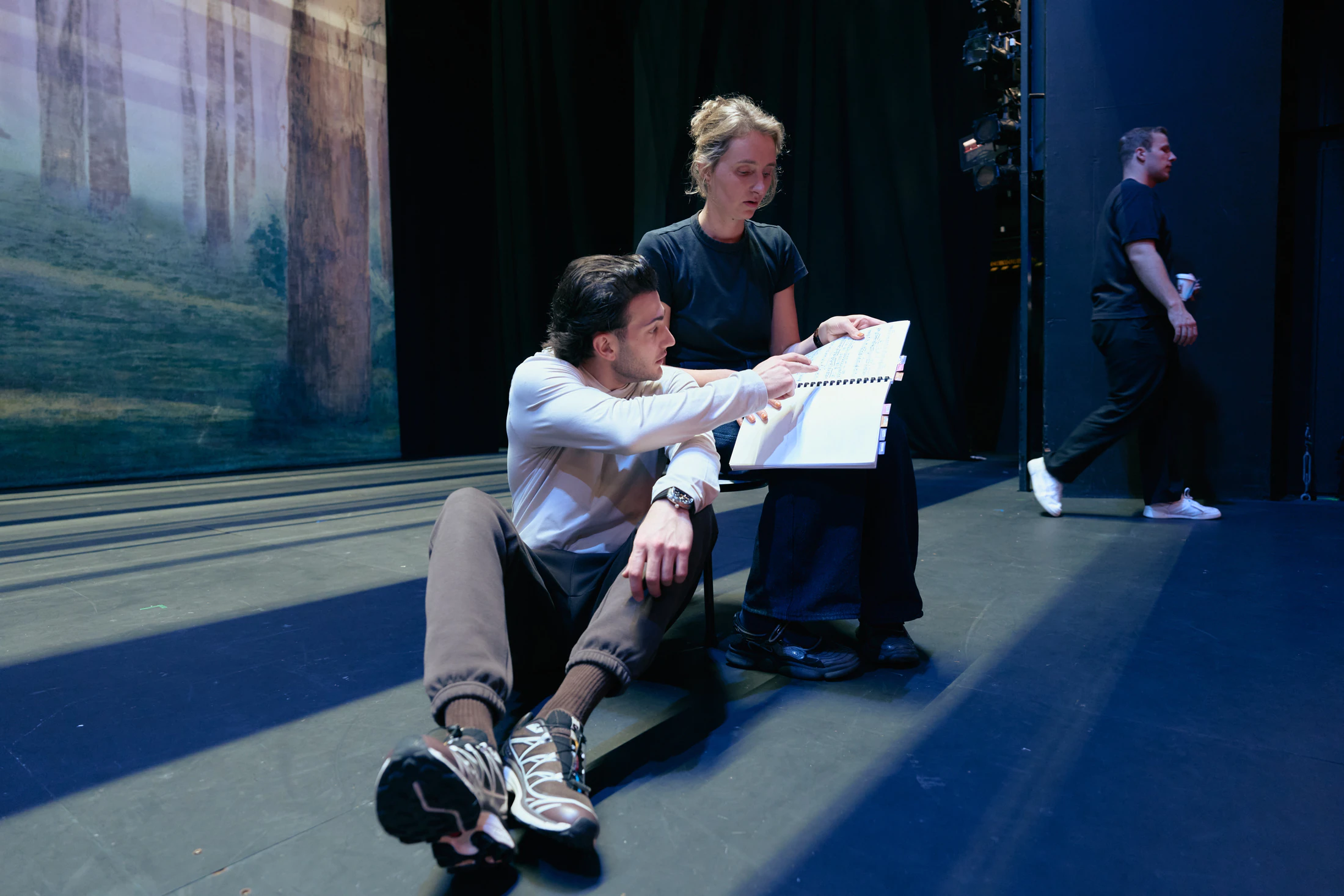 Two singers rehearse on the stage floor against a painted forest backdrop