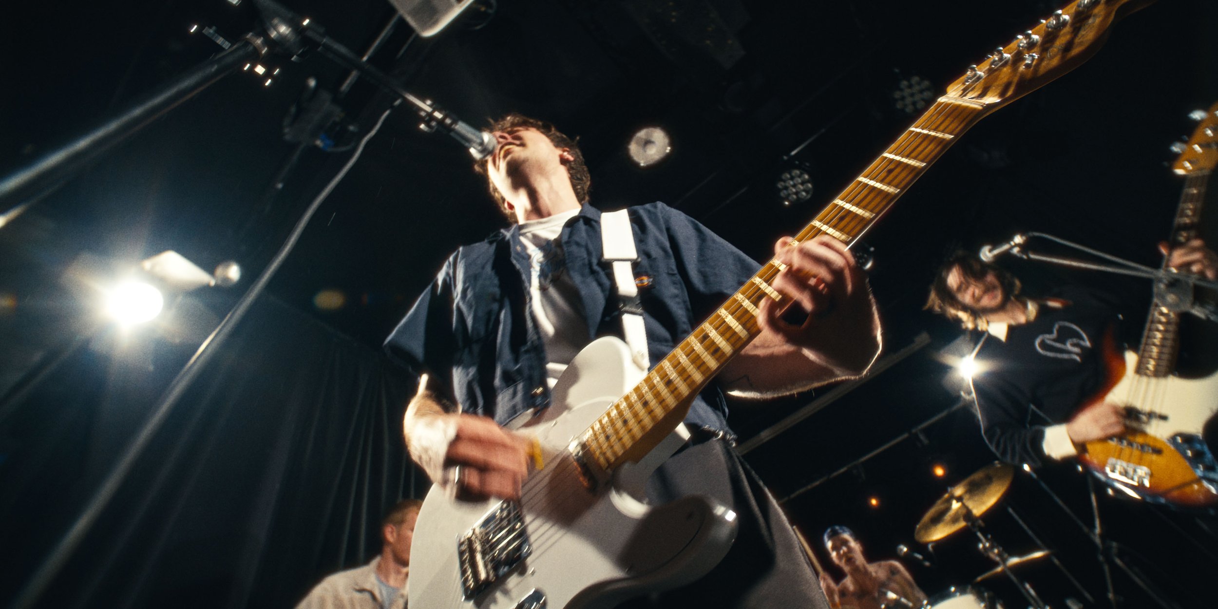 The singer leans into the mic over a white guitar, shot from below