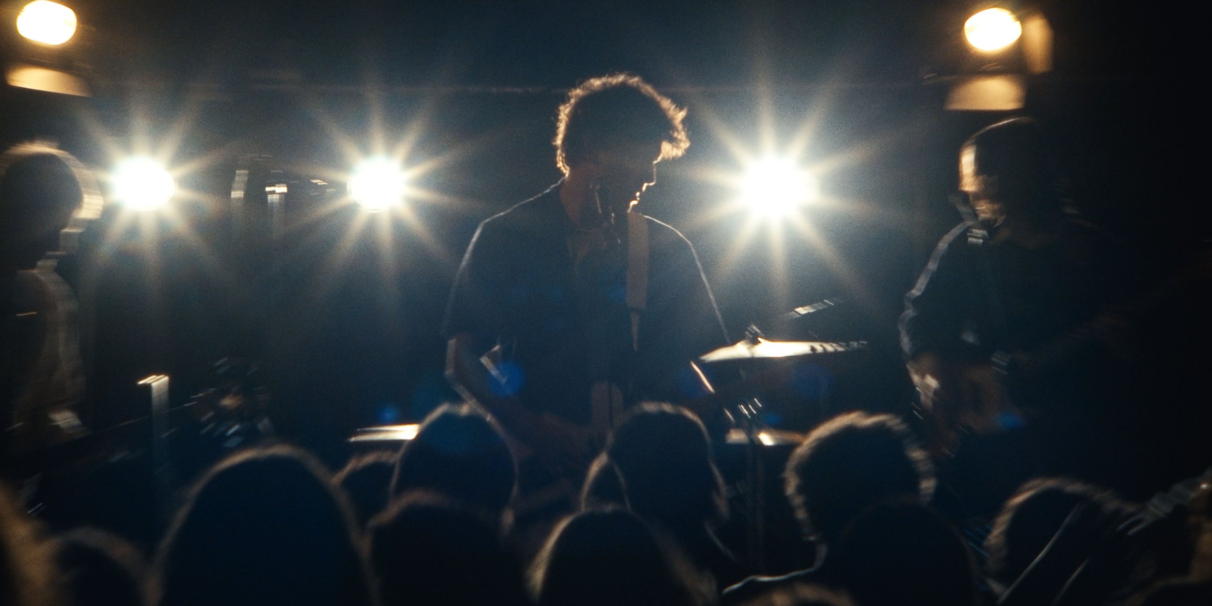 The guitarist through silhouetted heads in the crowd, stage lights flaring