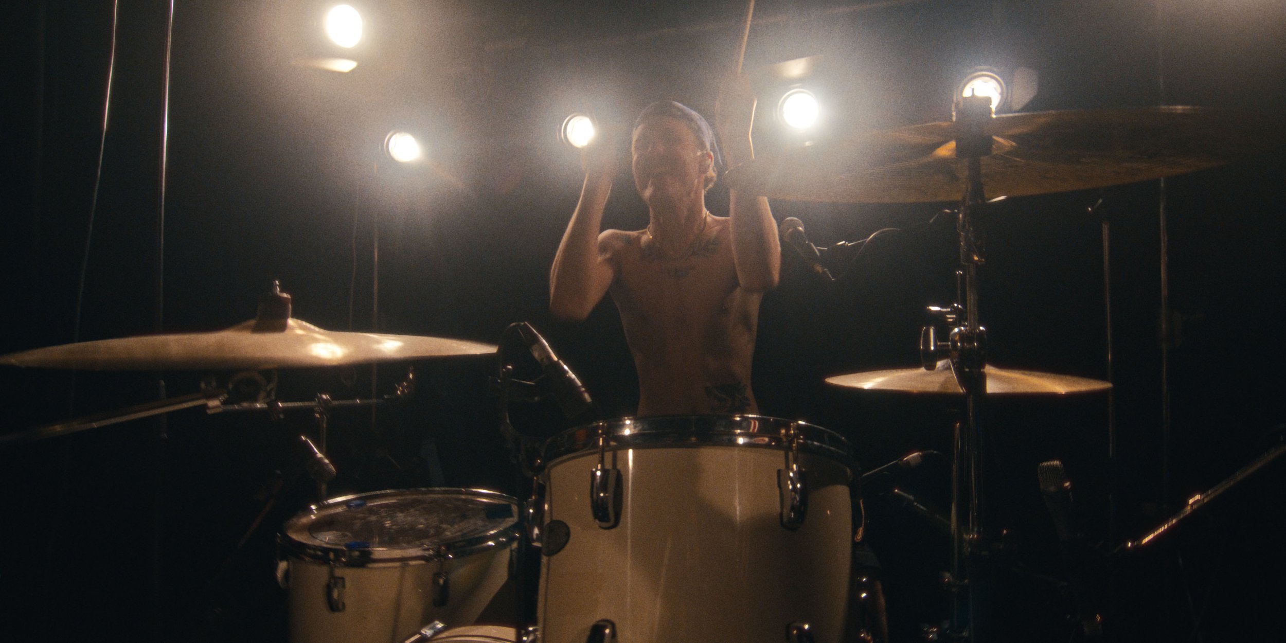 The drummer stands behind the kit, sticks raised, under warm lights