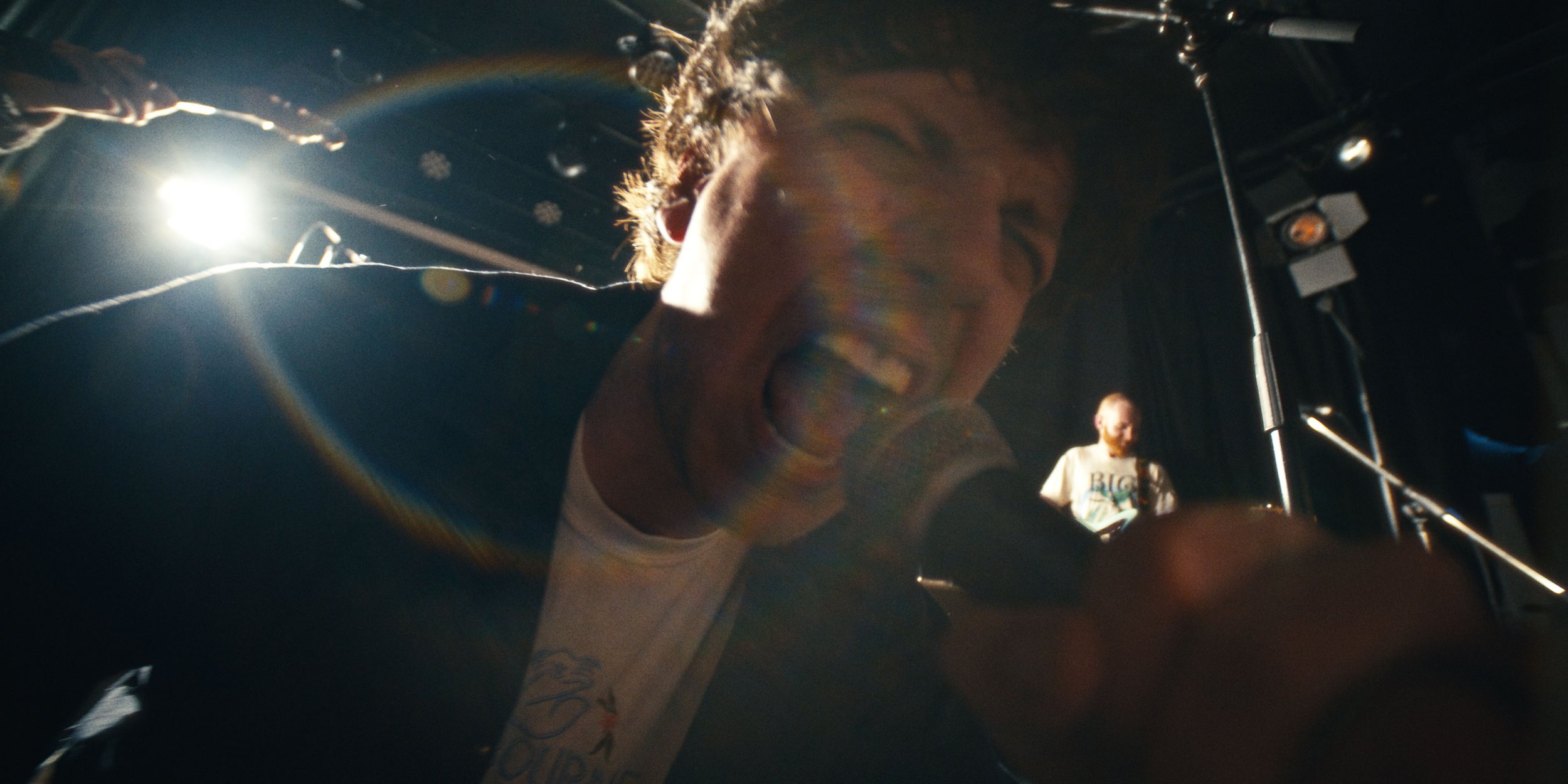 A singer screams into the mic close to the lens, a lens flare behind