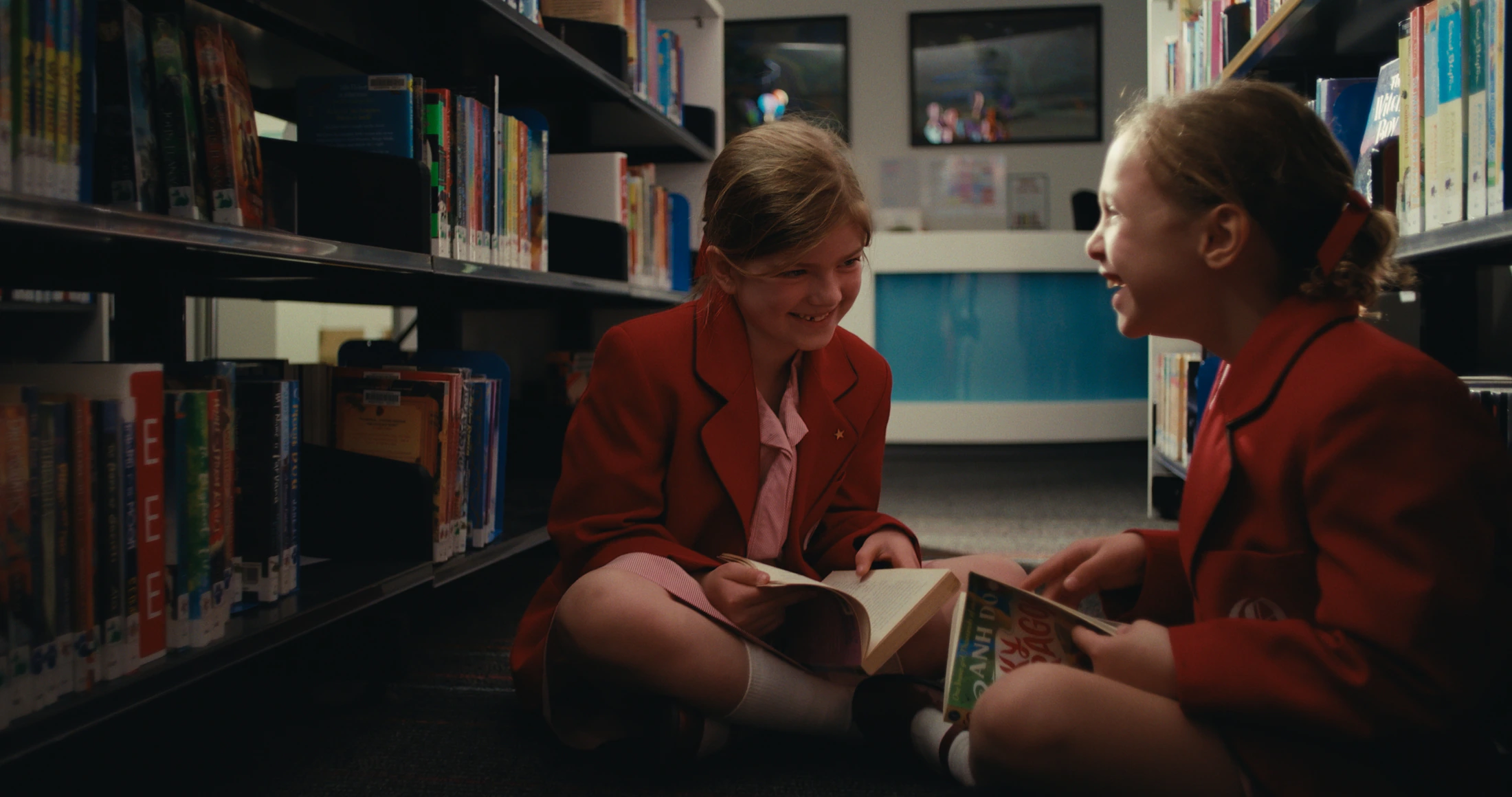 Two junior students in red blazers sit on the library floor laughing over a book