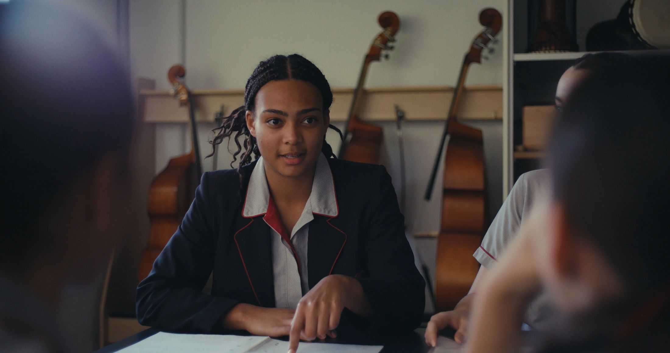 A senior student in a navy blazer looks up from her work in the music room, cellos behind her
