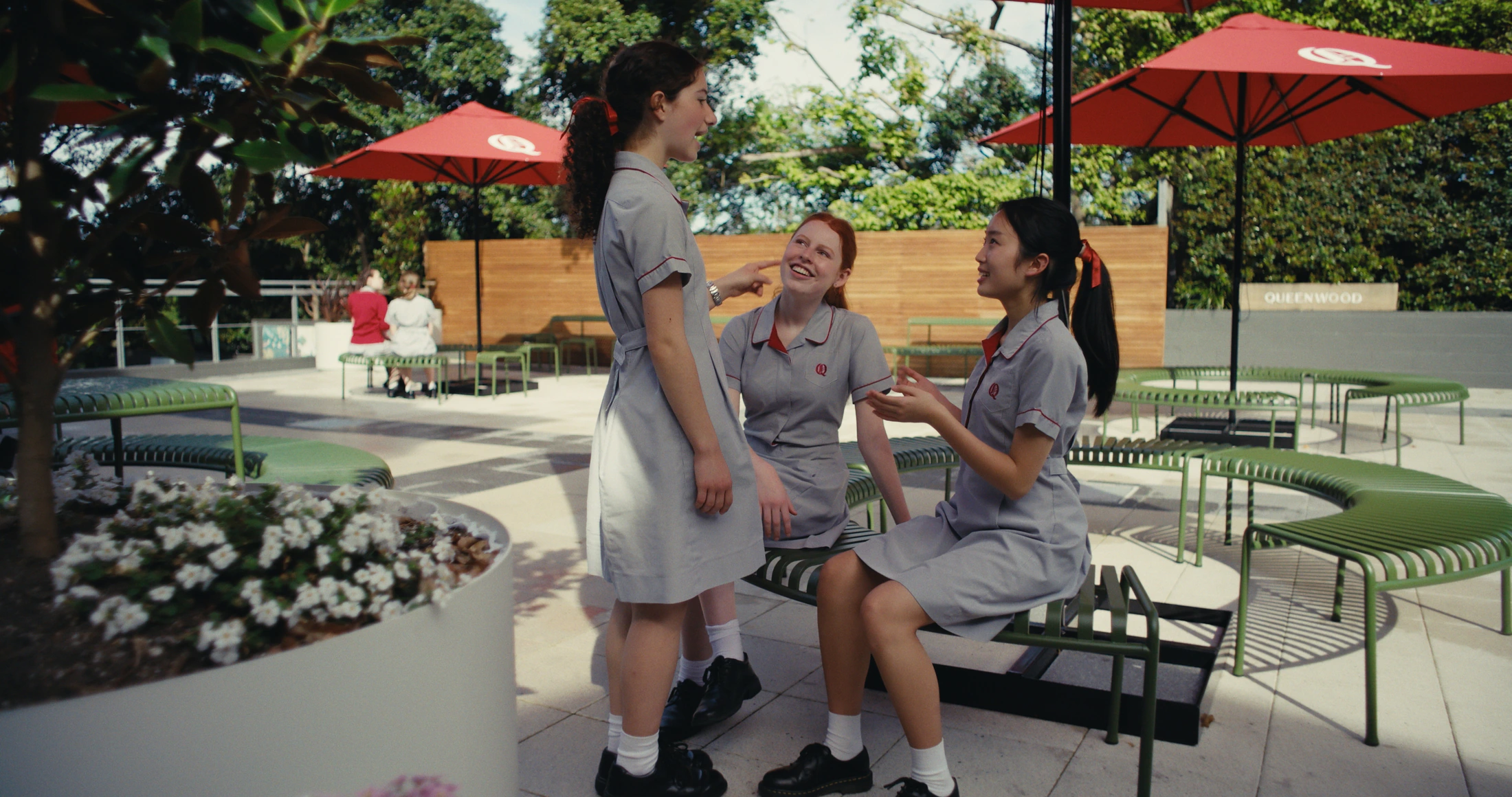 Three students talk and laugh on benches beneath red umbrellas in a courtyard