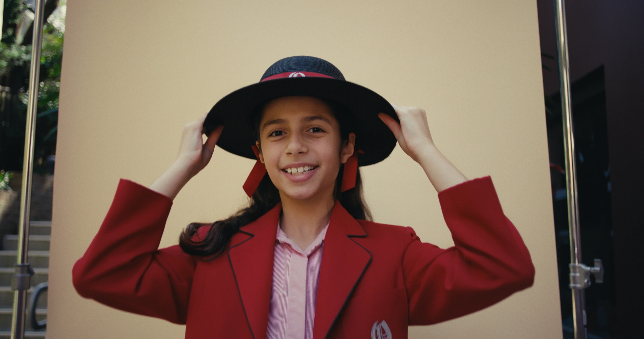 A student in a red blazer holds the brim of her boater and smiles