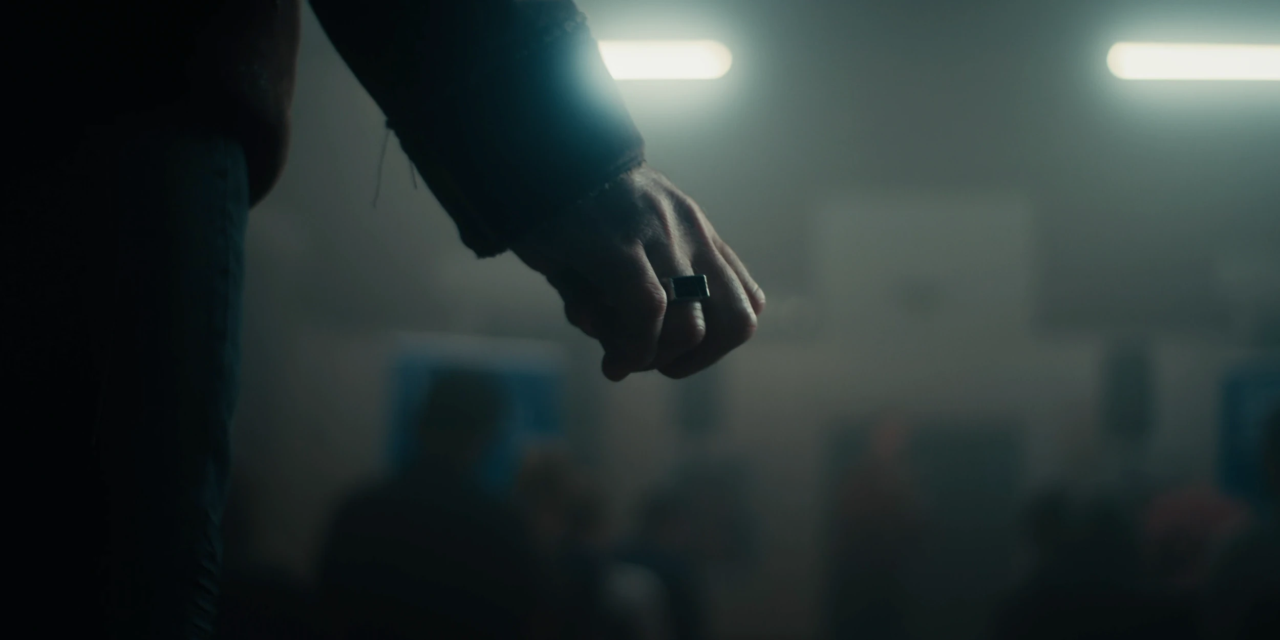 A clenched hand with a ring hangs at a man's side in a smoky hall under fluorescent lights