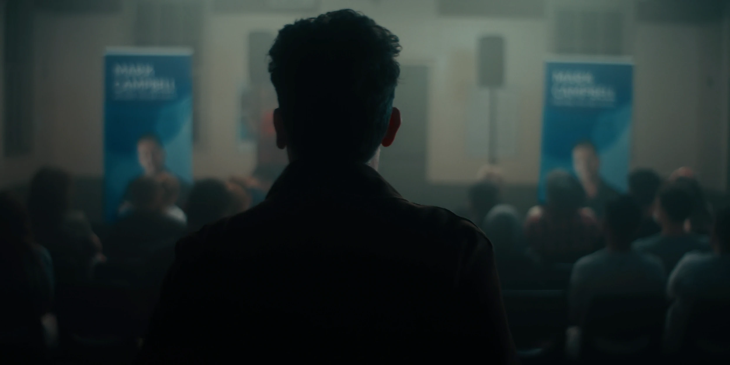 A man in silhouette faces a seated audience between campaign banners