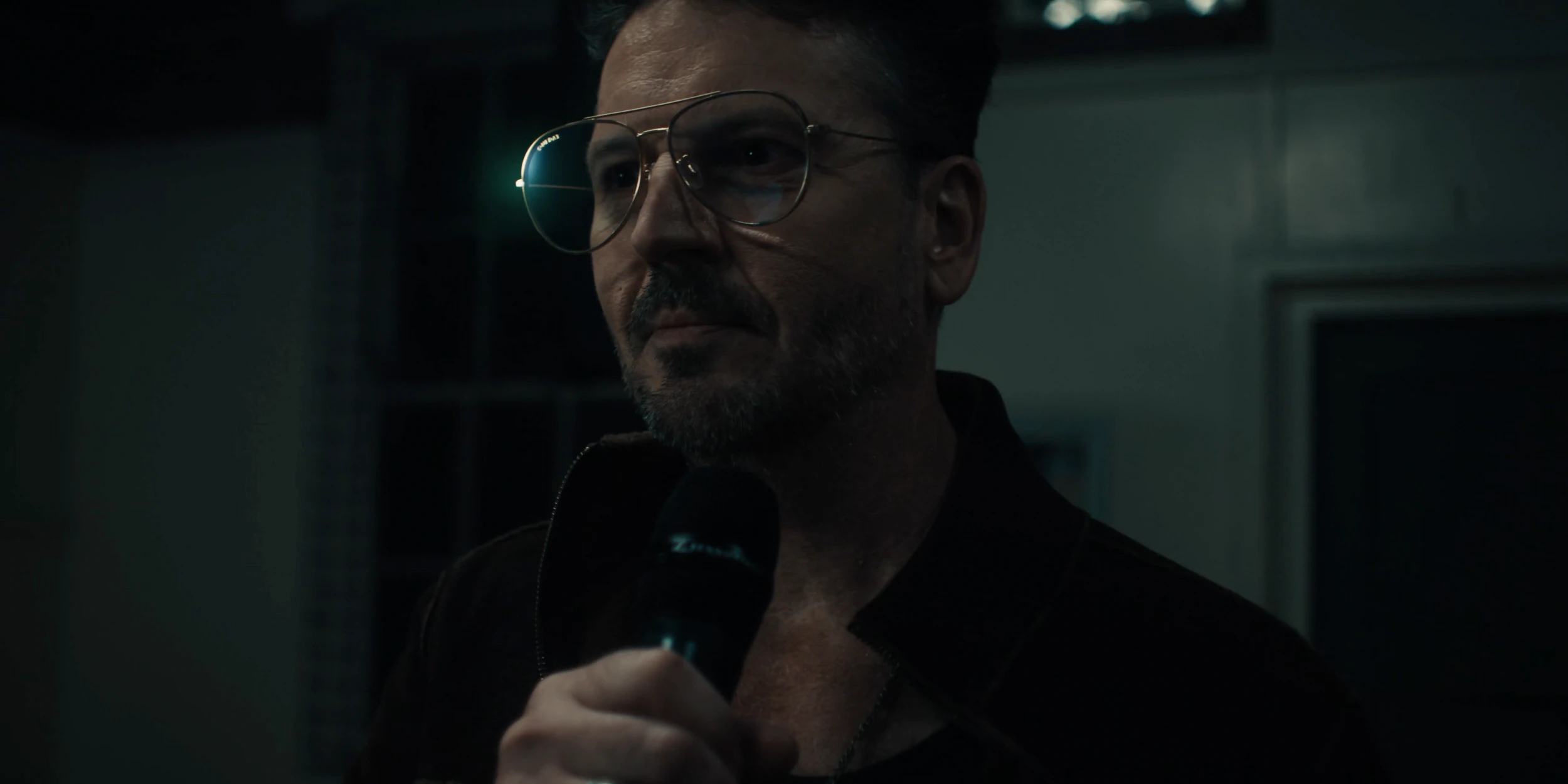 The speaker in aviator glasses holds a microphone, looking off