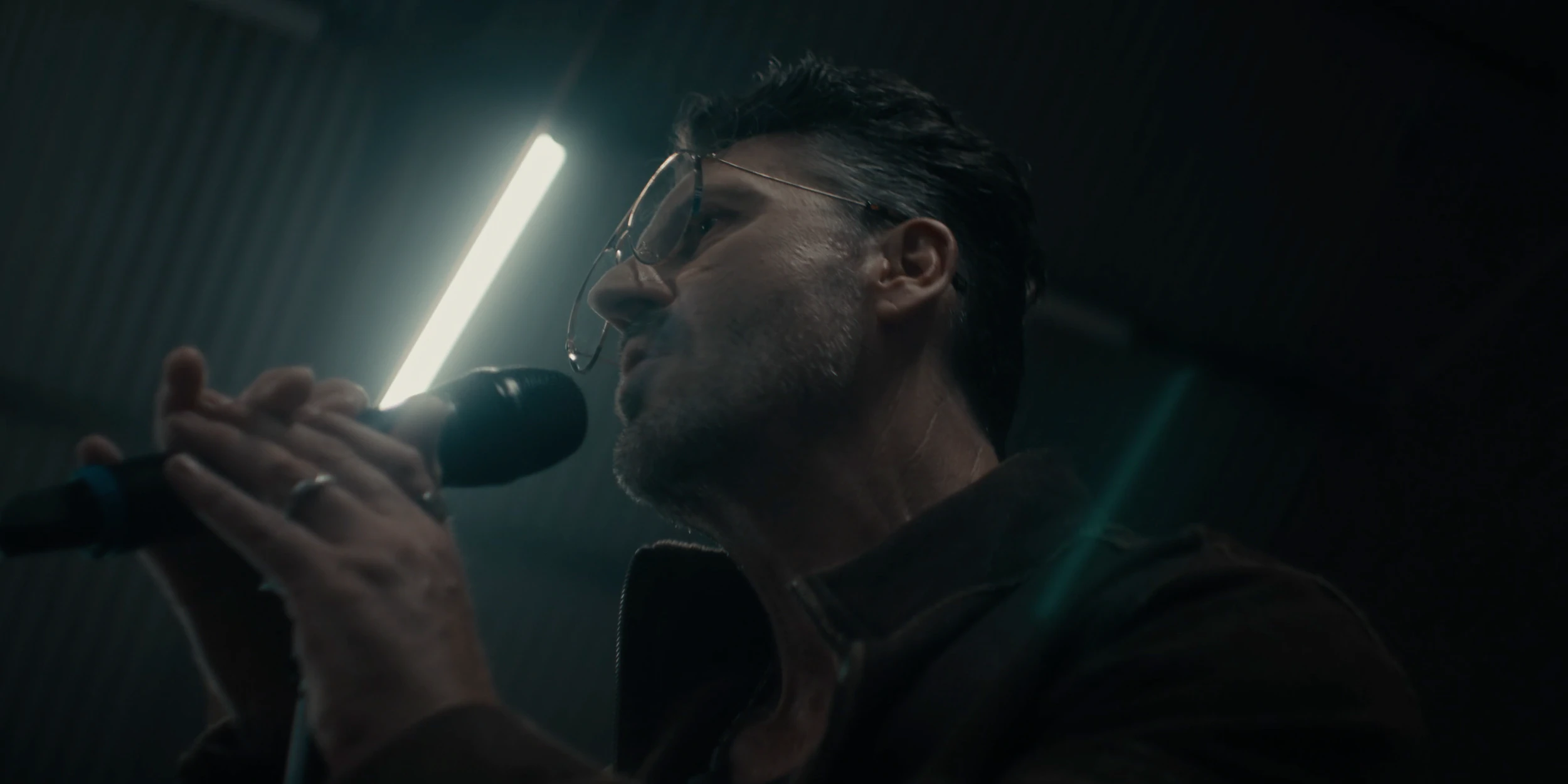 The speaker grips a microphone under a fluorescent tube, lit from the side