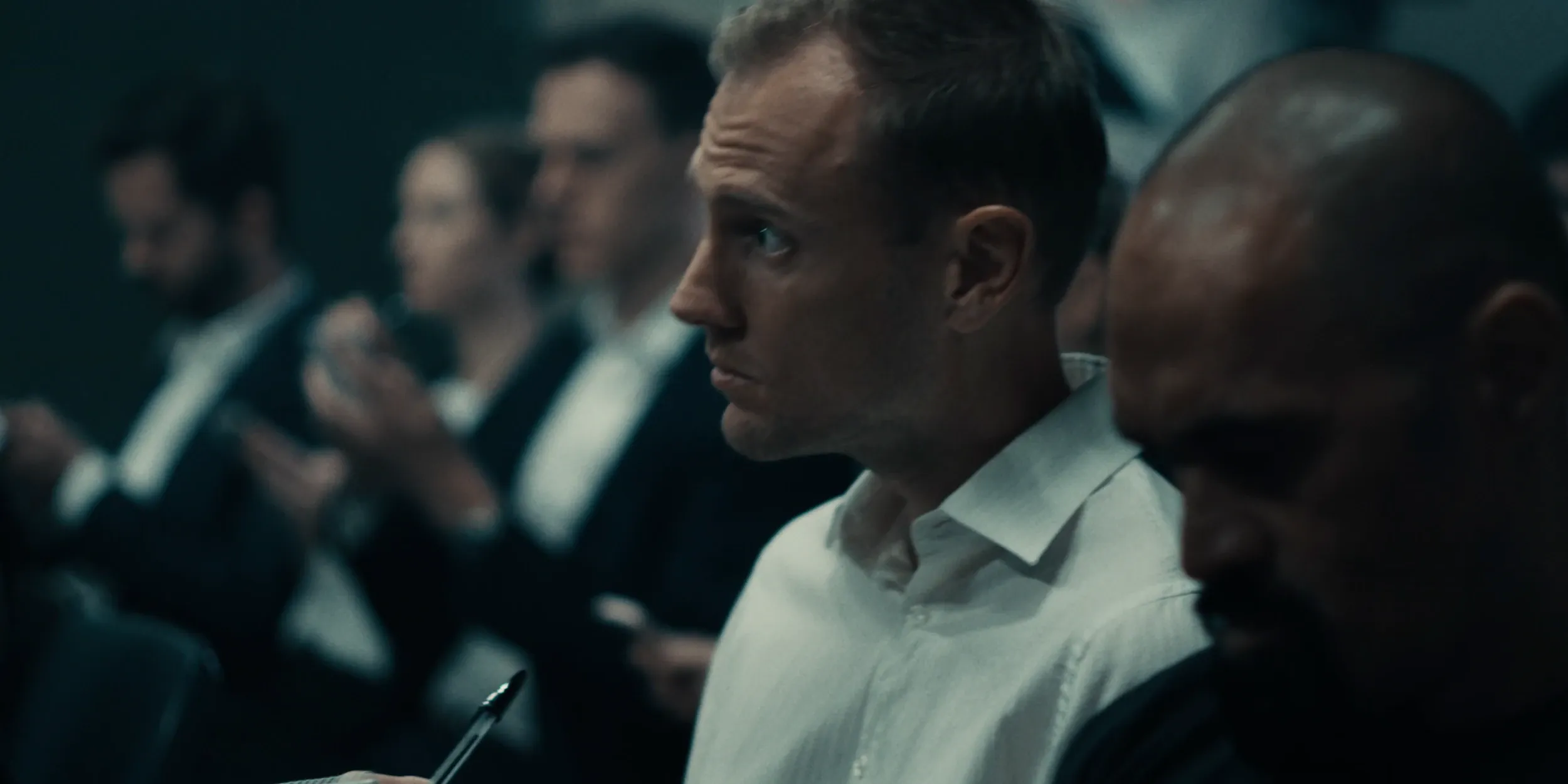 A man in a white shirt listens in a row of suited audience members